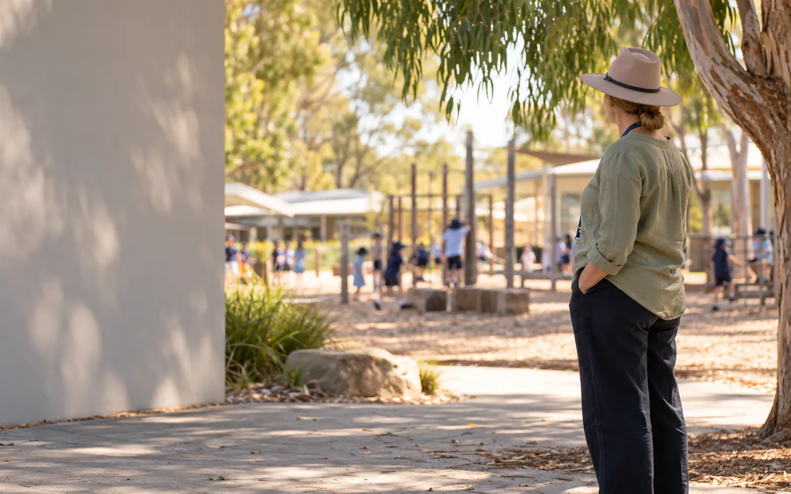 A teacher on yard duty in a hat and lanyard watching a school playground at lunchtime from a distance.