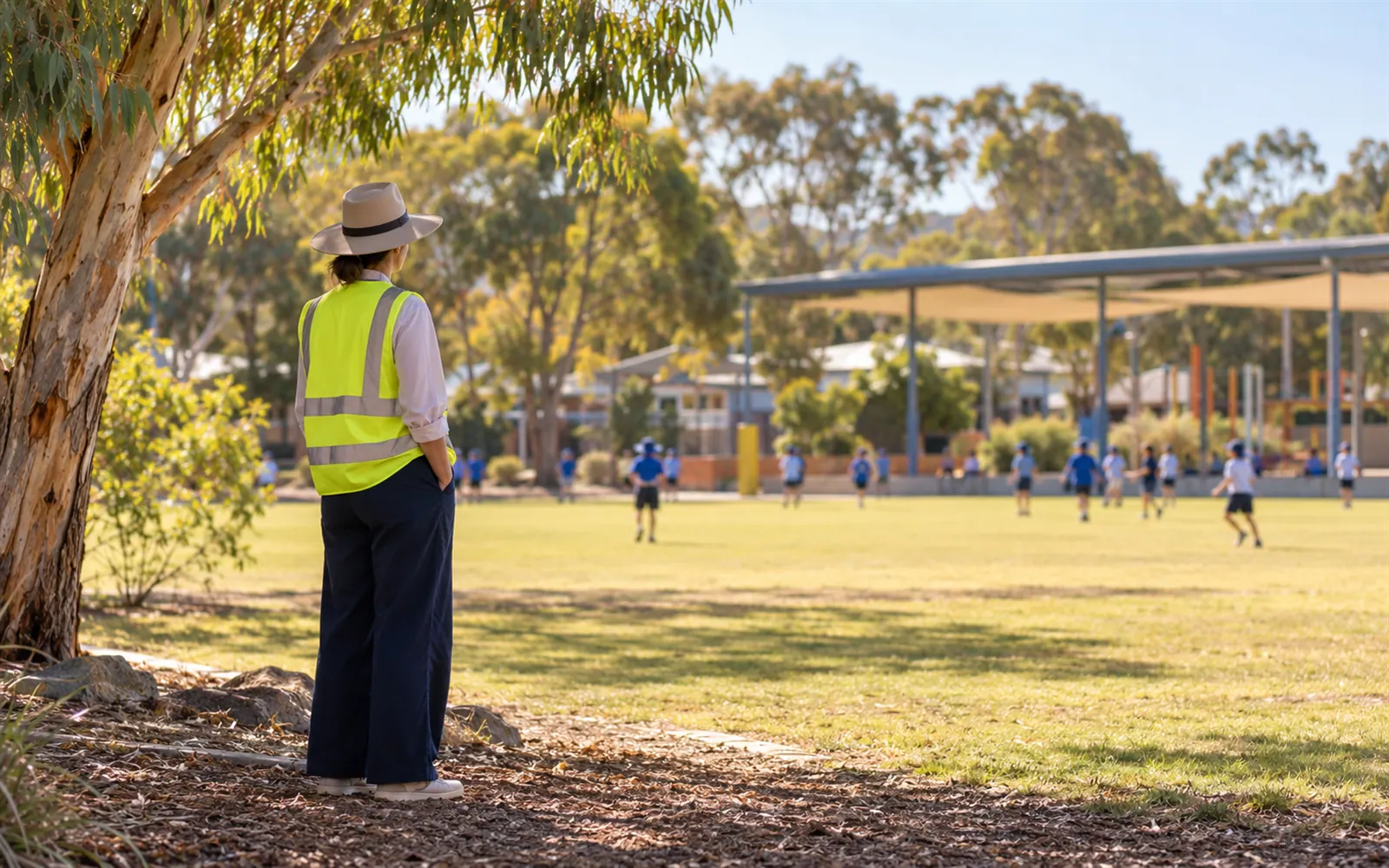A teacher in a hat and vest supervising the school oval at recess.