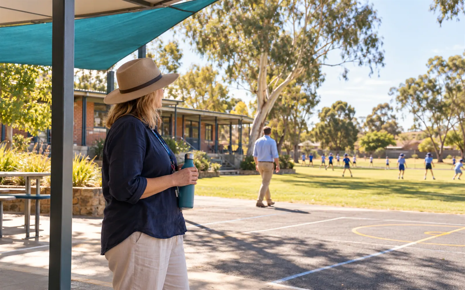 A teacher in a sun hat on yard duty watches a school playground at lunchtime while a colleague walks along the oval.